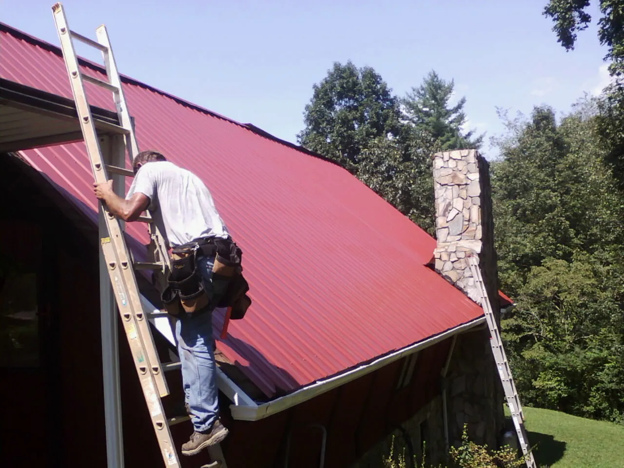 Roofer on a ladder beside a red metal roof and stone chimney — Soffit & Fascia Repair by Roofing Brothers in Diamond Bar